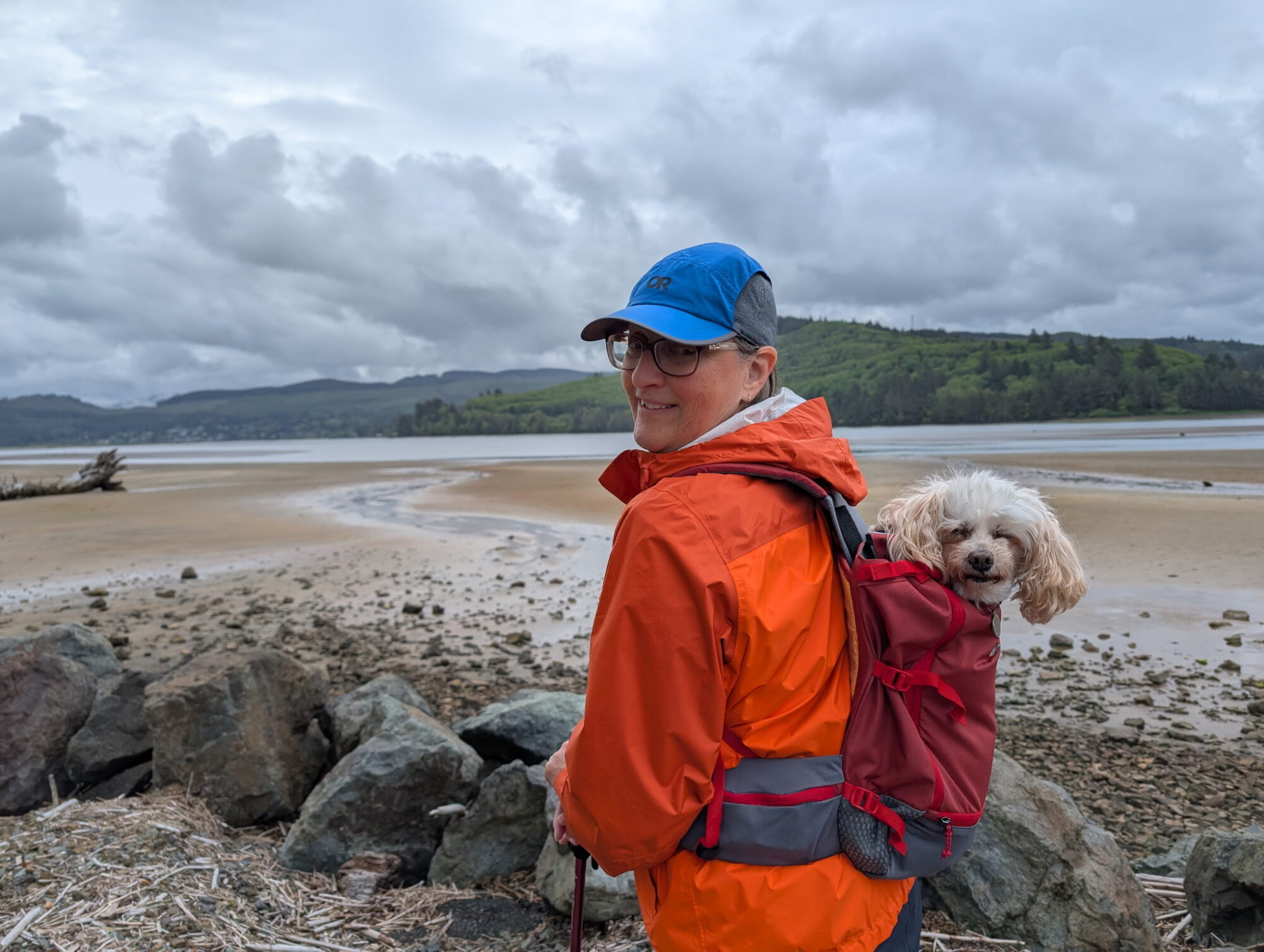 A women smiles at the camera with a small poodle in a backpack on her back. Behind her is a bay and some mountains