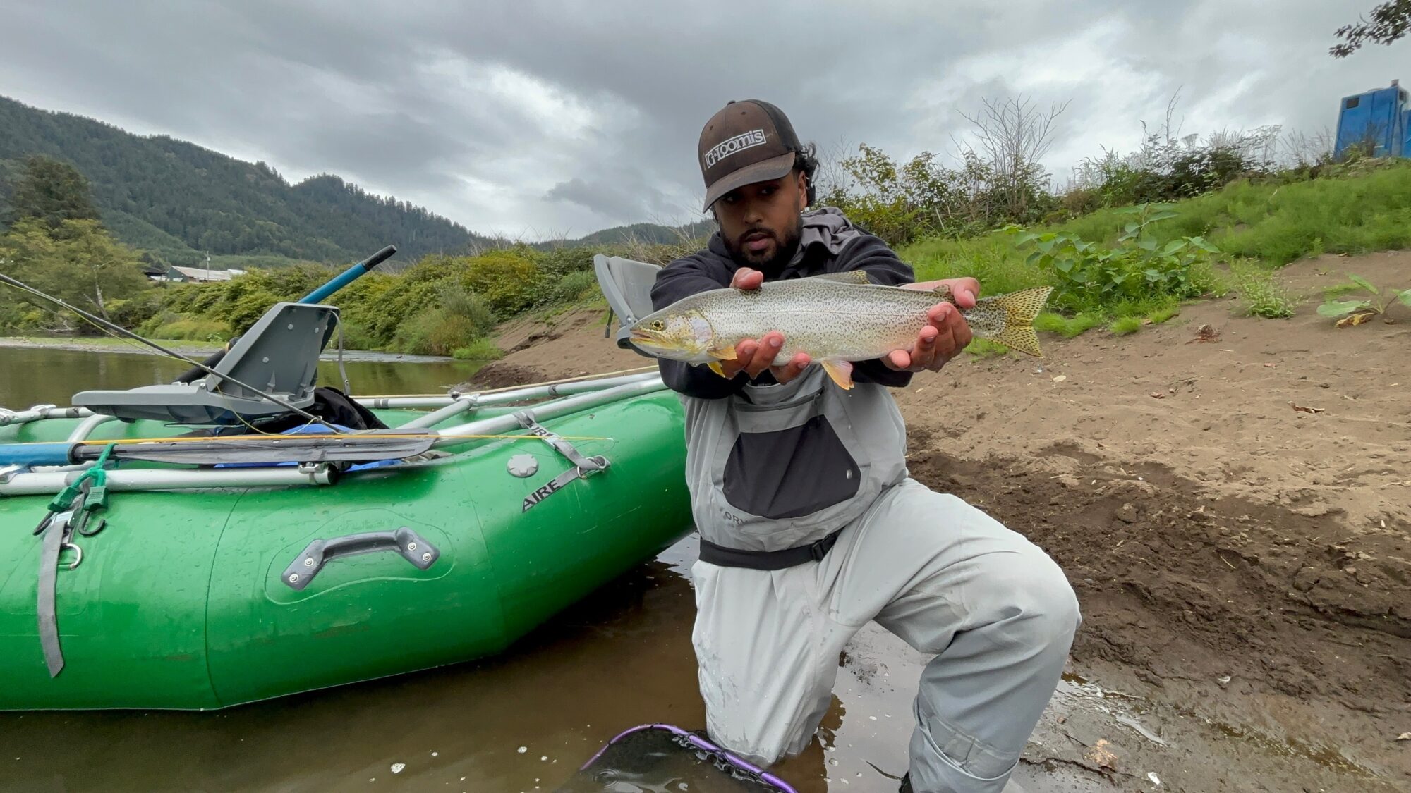 A man holds a cutthroat trout fish up to the camera behind him is a pontoon boat and the sandy bank of the river