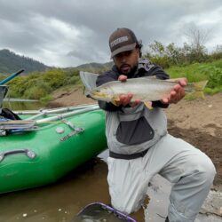 A man holds a cutthroat trout fish up to the camera behind him is a pontoon boat and the sandy bank of the river