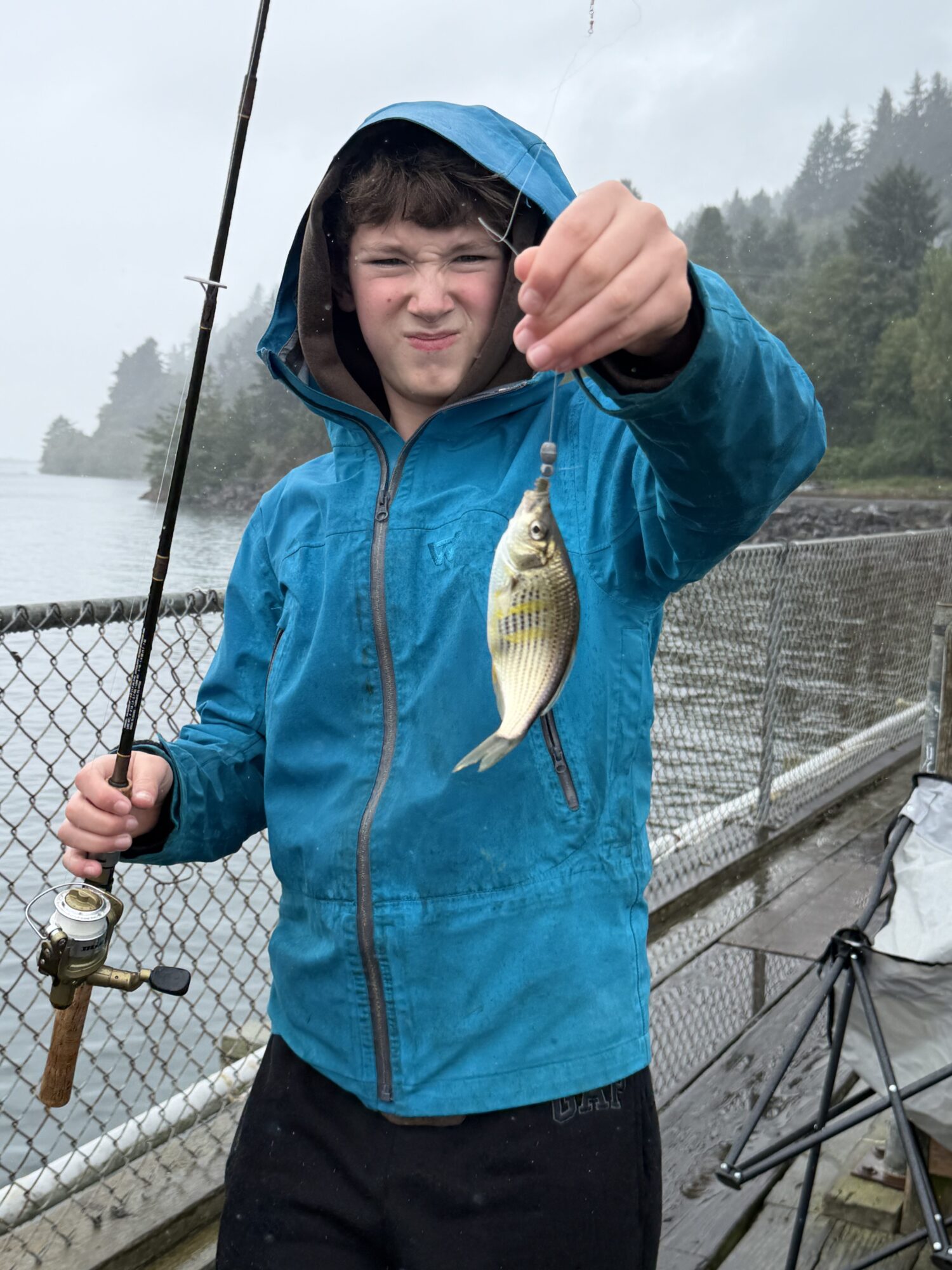 A boy in a blue jacket standing on a dock holding a fishing pole with a small surf perch caught on the end of the pole.