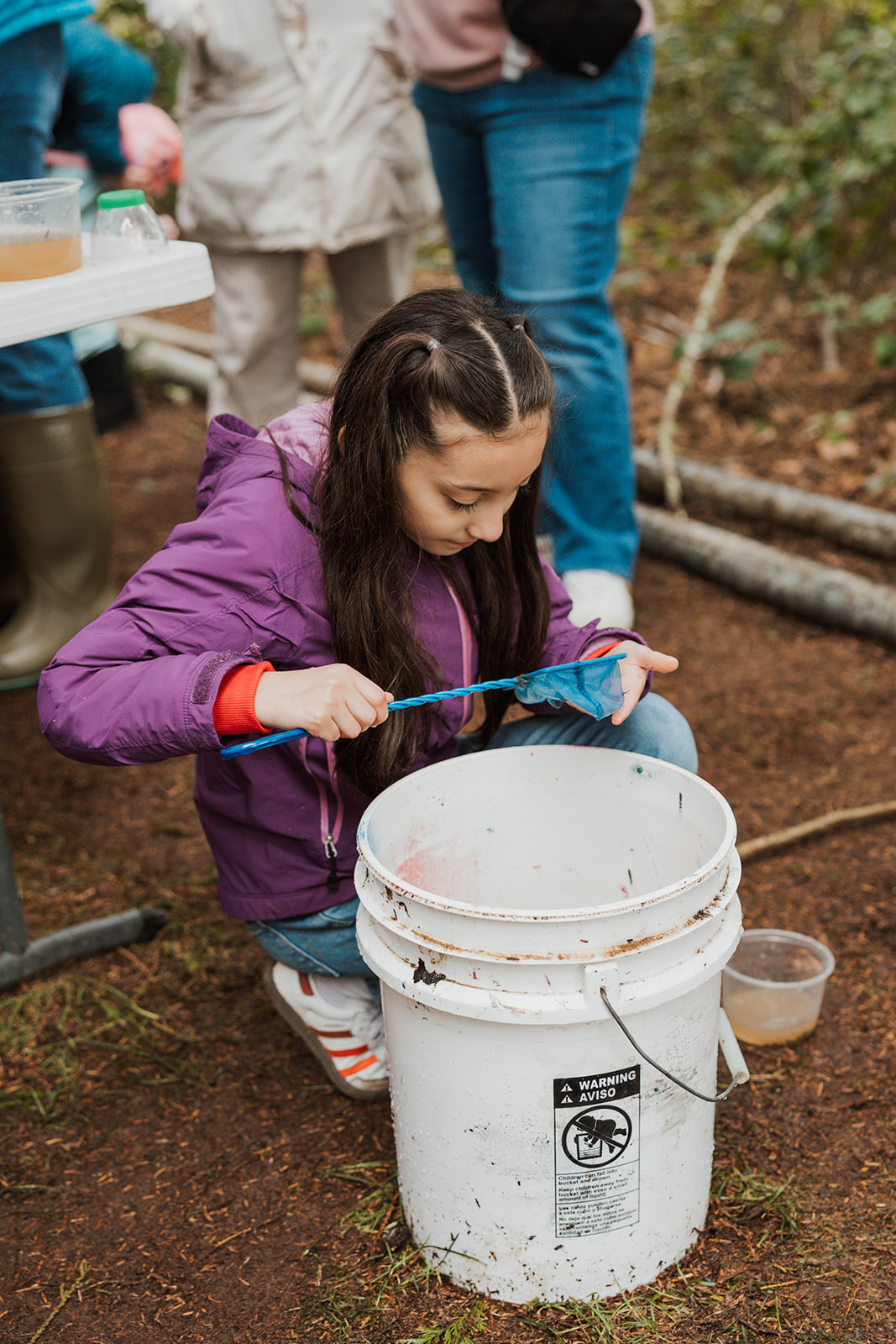 Girl in Pink Sweater holding small green net looking at something she scooped from a white bucket