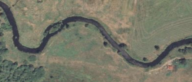 Aerial view of a winding stream surrounded by fields and minimal trees, with some buildings in the lower right corner.